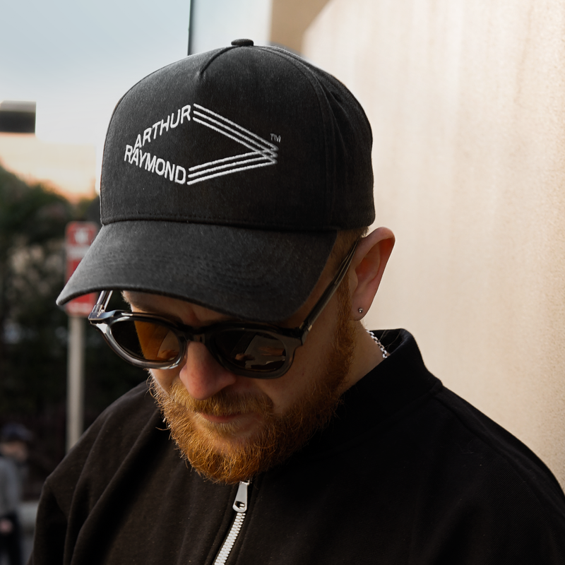 Arthur Raymond unisex vintage washed grey 5 panel baseball cap with peak showing white embroidered Arthur Raymond logo on front panel, modelled by man with red beard and sunglasses wearing Arthur Raymond men's Quarter Zip Bomber in black, looking down